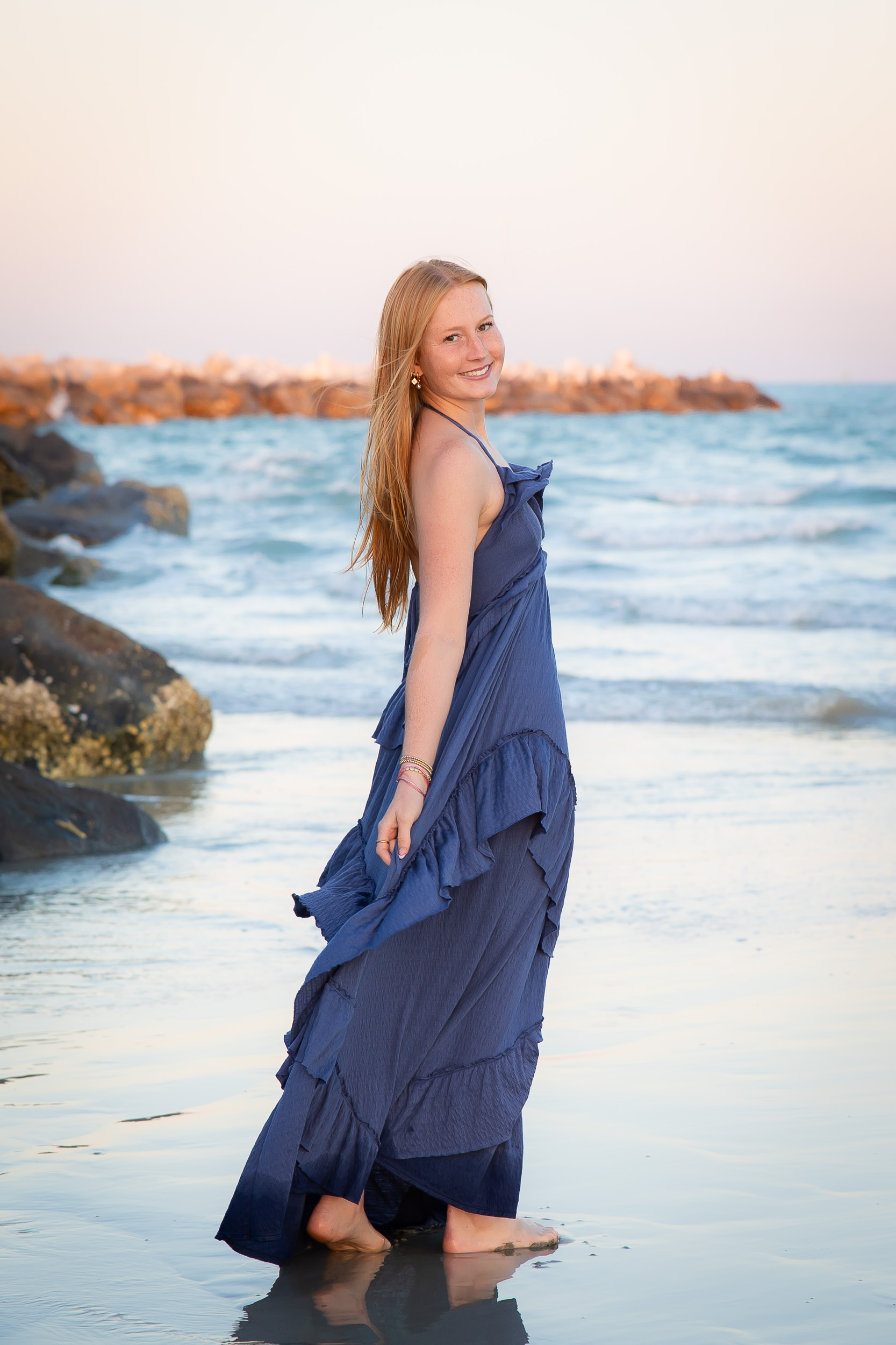 High school senior girl in a blue dress walking along the shoreline at Jetty Park in Cape Canaveral, Florida.