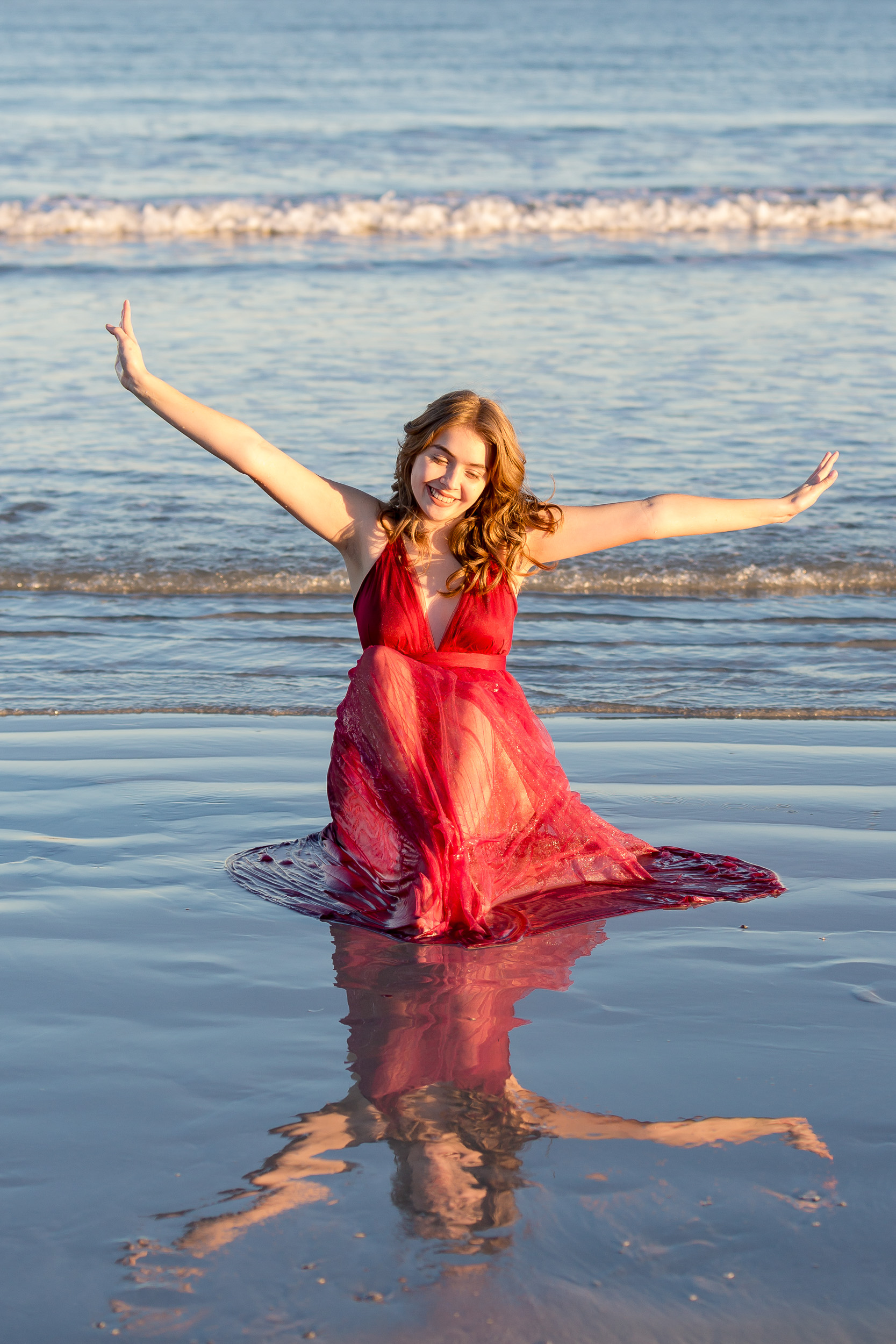 High school senior girl sitting in the ocean and splashing in her red dress during a senior photo session at Jetty Park in Cape Canaveral, Florida.